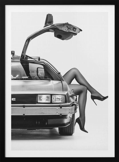 A black and white studio photograph of a DeLorean car with its gull-wing door open, featuring a woman's legs in black stockings and stilettos elegantly emerging from the passenger side. Decor