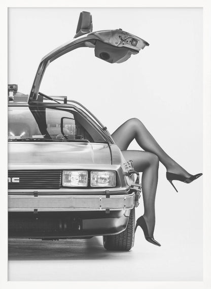 A black and white studio photograph of a DeLorean car with its gull-wing door open, featuring a woman's legs in black stockings and stilettos elegantly emerging from the passenger side. Decor