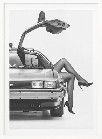 A black and white studio photograph of a DeLorean car with its gull-wing door open, featuring a woman's legs in black stockings and stilettos elegantly emerging from the passenger side. Decor