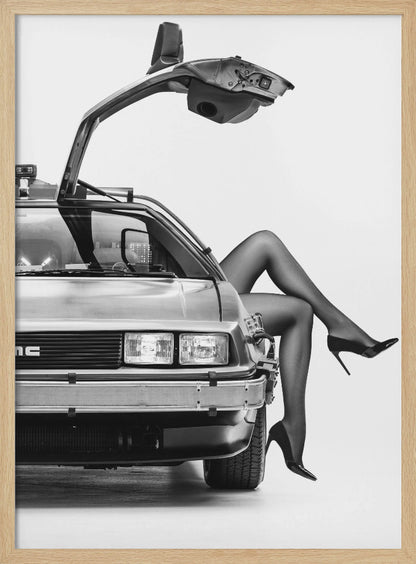 A black and white studio photograph of a DeLorean car with its gull-wing door open, featuring a woman's legs in black stockings and stilettos elegantly emerging from the passenger side. Decor