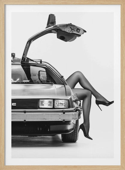 A black and white studio photograph of a DeLorean car with its gull-wing door open, featuring a woman's legs in black stockings and stilettos elegantly emerging from the passenger side. Decor