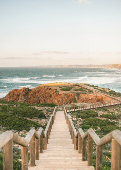 A wooden staircase and walkway descends a green, scrub-covered hill towards a rocky point overlooking a churning blue ocean under a pale, hazy sky. Print