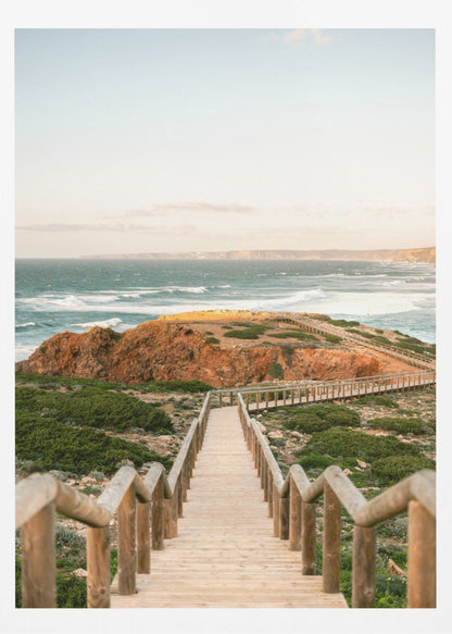 A wooden staircase and walkway descends a green, scrub-covered hill towards a rocky point overlooking a churning blue ocean under a pale, hazy sky. Print