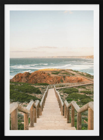 A wooden staircase and walkway descends a green, scrub-covered hill towards a rocky point overlooking a churning blue ocean under a pale, hazy sky. Print