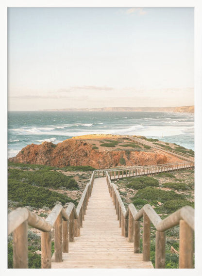 A wooden staircase and walkway descends a green, scrub-covered hill towards a rocky point overlooking a churning blue ocean under a pale, hazy sky. Print