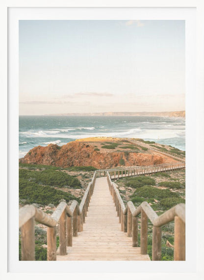 A wooden staircase and walkway descends a green, scrub-covered hill towards a rocky point overlooking a churning blue ocean under a pale, hazy sky. Print