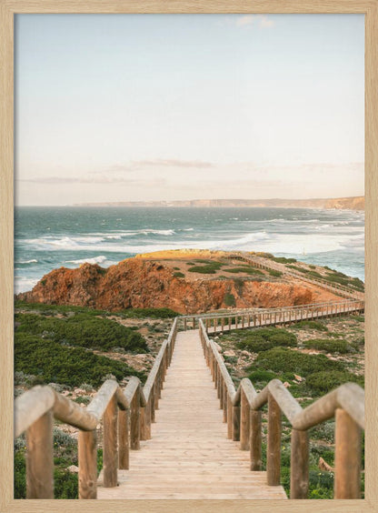 A wooden staircase and walkway descends a green, scrub-covered hill towards a rocky point overlooking a churning blue ocean under a pale, hazy sky. Print