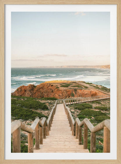 A wooden staircase and walkway descends a green, scrub-covered hill towards a rocky point overlooking a churning blue ocean under a pale, hazy sky. Print