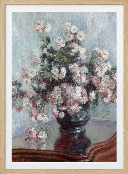 An impressionist painting by Claude Monet depicting a large bouquet of white and pink chrysanthemums in a dark, ornate vase. The vase sits on a reflective, reddish-brown wooden table, and the background is a textured light blue wall. The painting is framed in black. Artwork