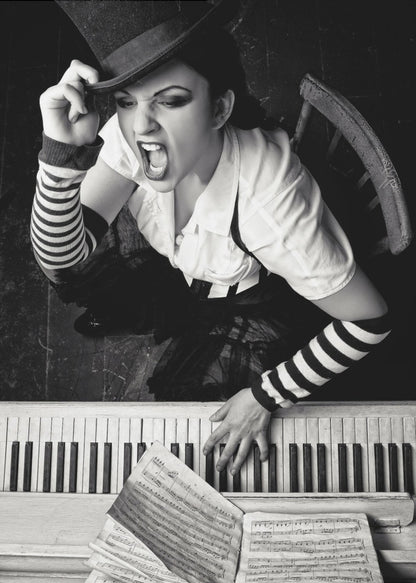 A dramatic black and white high-angle photograph of a woman in a top hat and striped arm warmers, shouting or singing passionately while seated at a piano with sheet music. Print