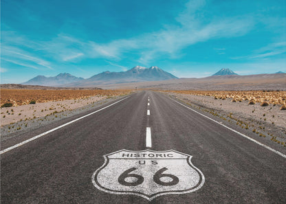 A framed photograph of the iconic Historic US Route 66 shield painted on a long, straight asphalt road that stretches through a desert landscape toward distant snow-capped mountains under a vibrant blue sky. Wall Art
