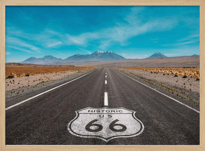 A framed photograph of the iconic Historic US Route 66 shield painted on a long, straight asphalt road that stretches through a desert landscape toward distant snow-capped mountains under a vibrant blue sky. Wall Art