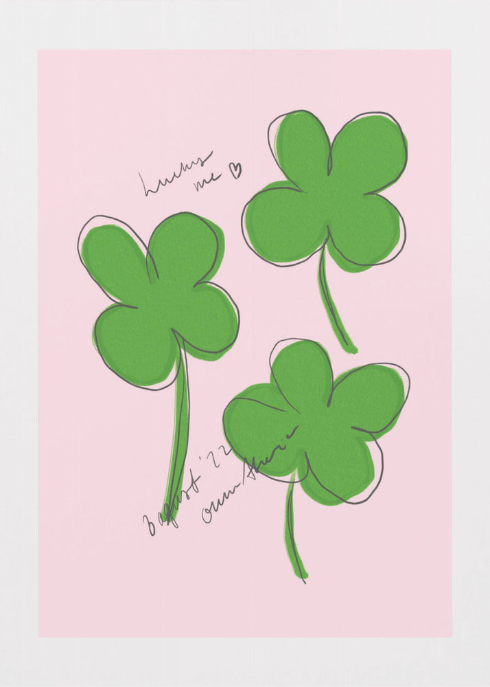 A framed art print featuring three hand-drawn four-leaf clovers in green with a loose black outline, set against a solid light pink background. Cursive handwriting on the print reads 'lucky me ♡' and includes a signature. Artwork