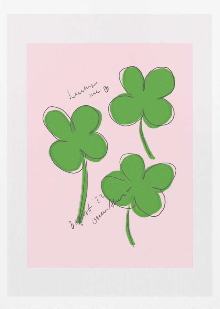 A framed art print featuring three hand-drawn four-leaf clovers in green with a loose black outline, set against a solid light pink background. Cursive handwriting on the print reads 'lucky me ♡' and includes a signature. Artwork