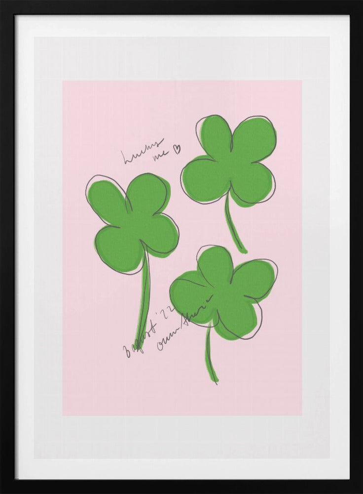 A framed art print featuring three hand-drawn four-leaf clovers in green with a loose black outline, set against a solid light pink background. Cursive handwriting on the print reads 'lucky me ♡' and includes a signature. Artwork