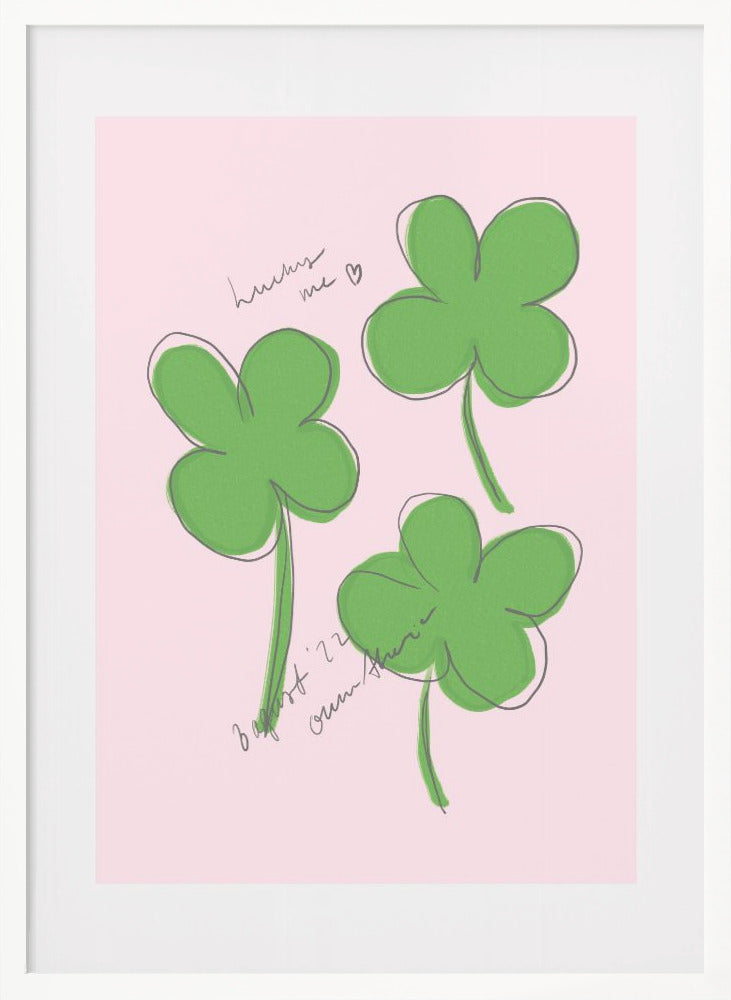A framed art print featuring three hand-drawn four-leaf clovers in green with a loose black outline, set against a solid light pink background. Cursive handwriting on the print reads 'lucky me ♡' and includes a signature. Artwork