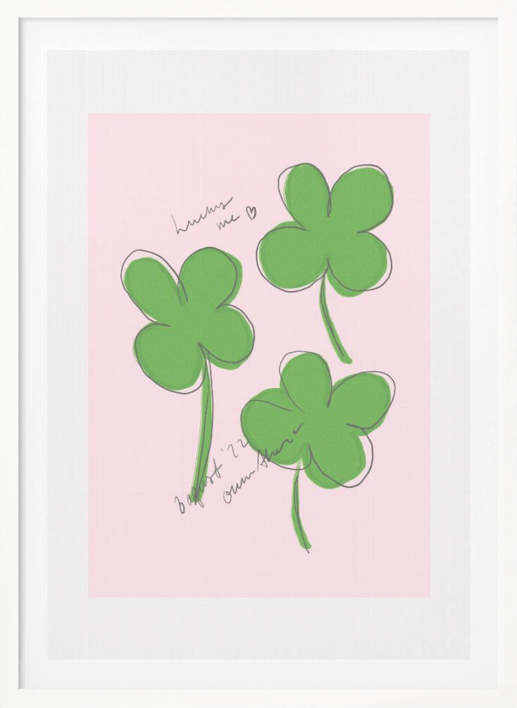 A framed art print featuring three hand-drawn four-leaf clovers in green with a loose black outline, set against a solid light pink background. Cursive handwriting on the print reads 'lucky me ♡' and includes a signature. Artwork