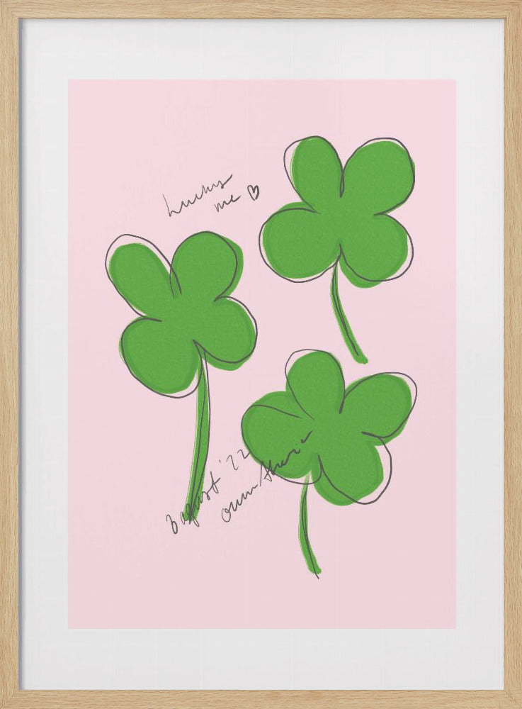 A framed art print featuring three hand-drawn four-leaf clovers in green with a loose black outline, set against a solid light pink background. Cursive handwriting on the print reads 'lucky me ♡' and includes a signature. Artwork
