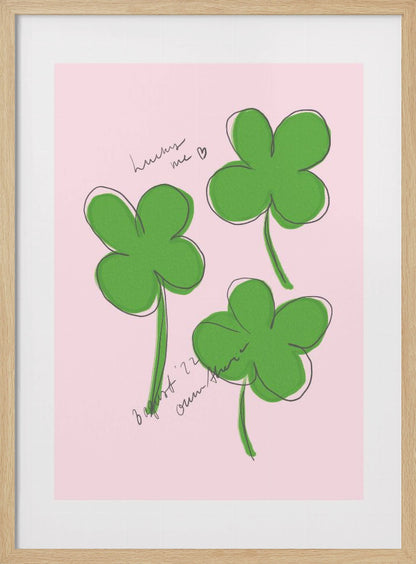 A framed art print featuring three hand-drawn four-leaf clovers in green with a loose black outline, set against a solid light pink background. Cursive handwriting on the print reads 'lucky me ♡' and includes a signature. Artwork