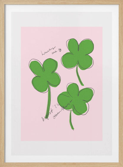 A framed art print featuring three hand-drawn four-leaf clovers in green with a loose black outline, set against a solid light pink background. Cursive handwriting on the print reads 'lucky me ♡' and includes a signature. Artwork