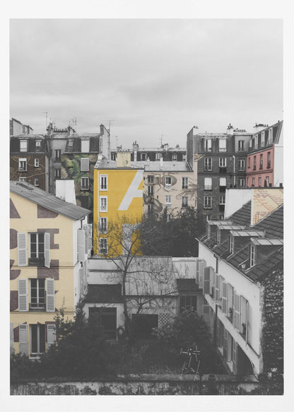 A framed, vertical photograph with selective color, showing a dense cluster of Parisian-style buildings under an overcast sky. Most of the image is in black and white, but one building is a vibrant yellow with a large white 'A' painted on it, creating a striking focal point. Wall Art