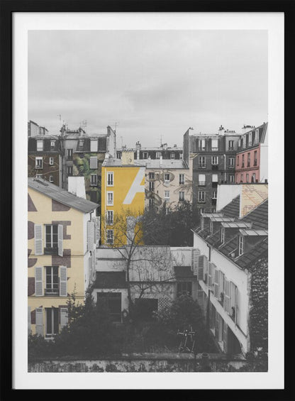 A framed, vertical photograph with selective color, showing a dense cluster of Parisian-style buildings under an overcast sky. Most of the image is in black and white, but one building is a vibrant yellow with a large white 'A' painted on it, creating a striking focal point. Wall Art