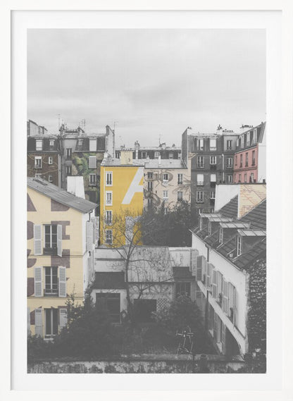 A framed, vertical photograph with selective color, showing a dense cluster of Parisian-style buildings under an overcast sky. Most of the image is in black and white, but one building is a vibrant yellow with a large white 'A' painted on it, creating a striking focal point. Wall Art