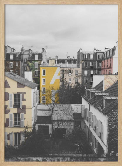 A framed, vertical photograph with selective color, showing a dense cluster of Parisian-style buildings under an overcast sky. Most of the image is in black and white, but one building is a vibrant yellow with a large white 'A' painted on it, creating a striking focal point. Wall Art