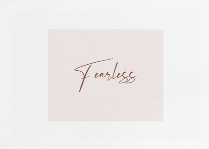 A framed artwork featuring the word 'Fearless' in a flowing brown script font on a pale pink background. The piece is surrounded by a white mat and a textured silver frame. Wall Art
