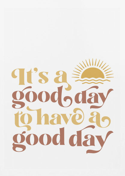 A framed art print with a white background featuring the quote 'It's a good day to have a good day' in a retro, curvy font. The text color alternates between terracotta brown and mustard yellow. Above the text is a small yellow icon of a sun rising over waves. Wall Art