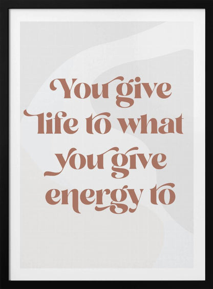 A framed motivational poster featuring the quote 'You give life to what you give energy to'. The text is printed in a dusty rose color using a stylish, retro serif font on a light gray background with subtle wavy shapes. Poster