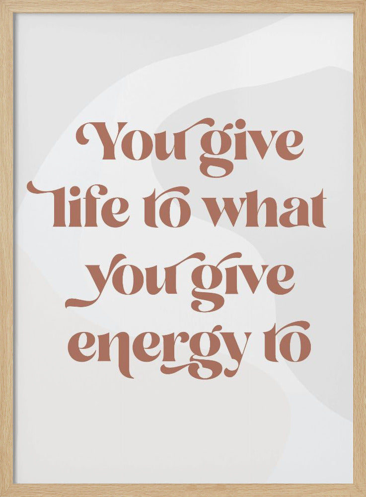 A framed motivational poster featuring the quote 'You give life to what you give energy to'. The text is printed in a dusty rose color using a stylish, retro serif font on a light gray background with subtle wavy shapes. Poster