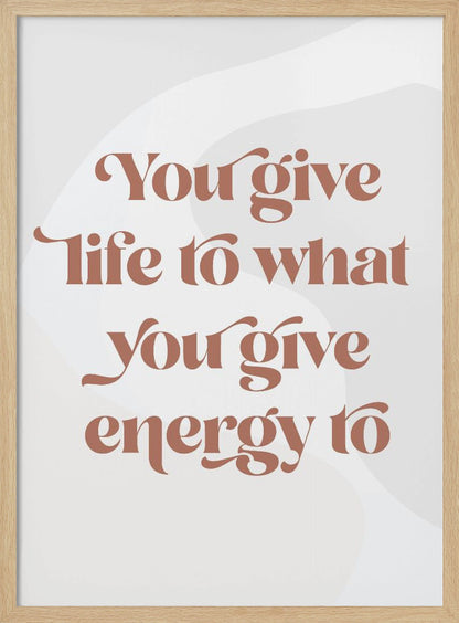 A framed motivational poster featuring the quote 'You give life to what you give energy to'. The text is printed in a dusty rose color using a stylish, retro serif font on a light gray background with subtle wavy shapes. Poster
