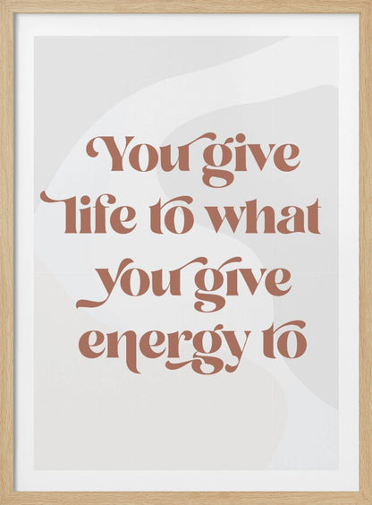 A framed motivational poster featuring the quote 'You give life to what you give energy to'. The text is printed in a dusty rose color using a stylish, retro serif font on a light gray background with subtle wavy shapes. Poster