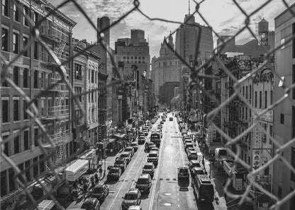A high-angle, black and white photograph of a bustling New York City street viewed through a chain-link fence. The fence is in the foreground, framing the scene of traffic-filled streets, historic buildings with fire escapes, and skyscrapers in the distance under a cloudy sky. Wall Art