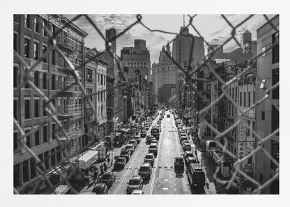 A high-angle, black and white photograph of a bustling New York City street viewed through a chain-link fence. The fence is in the foreground, framing the scene of traffic-filled streets, historic buildings with fire escapes, and skyscrapers in the distance under a cloudy sky. Wall Art