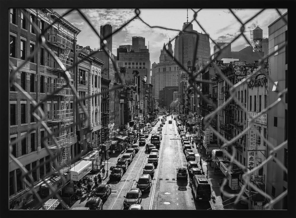 A high-angle, black and white photograph of a bustling New York City street viewed through a chain-link fence. The fence is in the foreground, framing the scene of traffic-filled streets, historic buildings with fire escapes, and skyscrapers in the distance under a cloudy sky. Wall Art