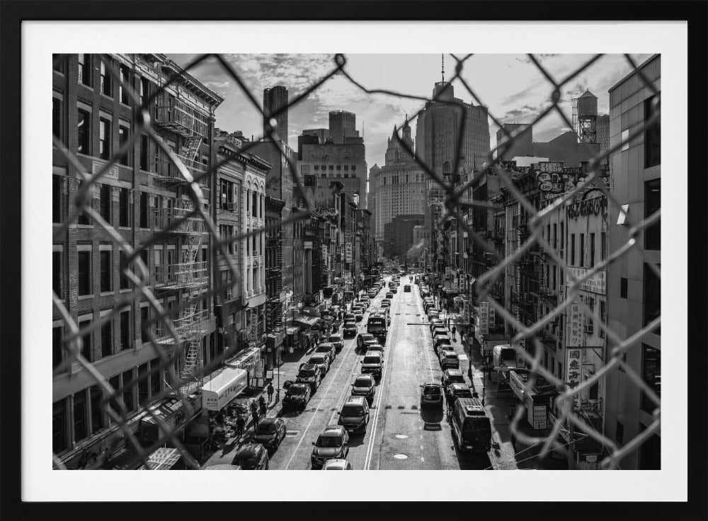 A high-angle, black and white photograph of a bustling New York City street viewed through a chain-link fence. The fence is in the foreground, framing the scene of traffic-filled streets, historic buildings with fire escapes, and skyscrapers in the distance under a cloudy sky. Wall Art