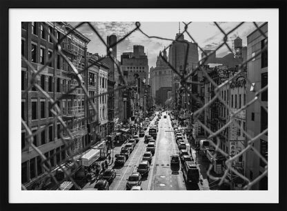 A high-angle, black and white photograph of a bustling New York City street viewed through a chain-link fence. The fence is in the foreground, framing the scene of traffic-filled streets, historic buildings with fire escapes, and skyscrapers in the distance under a cloudy sky. Wall Art