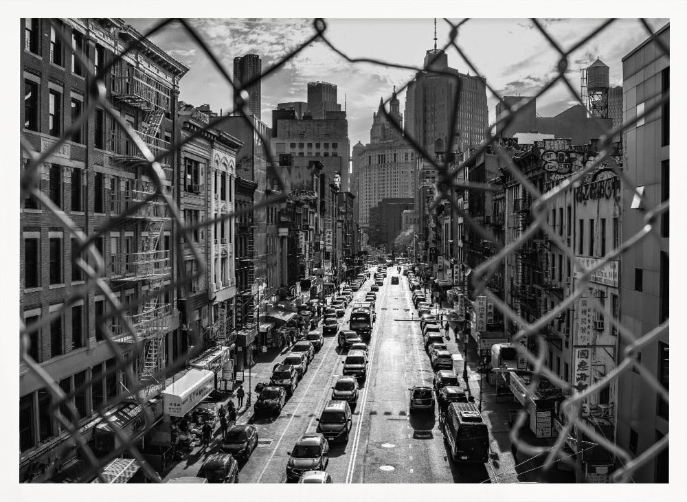 A high-angle, black and white photograph of a bustling New York City street viewed through a chain-link fence. The fence is in the foreground, framing the scene of traffic-filled streets, historic buildings with fire escapes, and skyscrapers in the distance under a cloudy sky. Wall Art