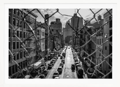 A high-angle, black and white photograph of a bustling New York City street viewed through a chain-link fence. The fence is in the foreground, framing the scene of traffic-filled streets, historic buildings with fire escapes, and skyscrapers in the distance under a cloudy sky. Wall Art