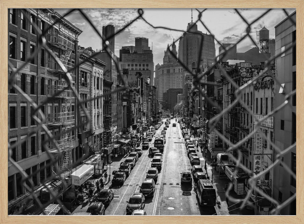 A high-angle, black and white photograph of a bustling New York City street viewed through a chain-link fence. The fence is in the foreground, framing the scene of traffic-filled streets, historic buildings with fire escapes, and skyscrapers in the distance under a cloudy sky. Wall Art