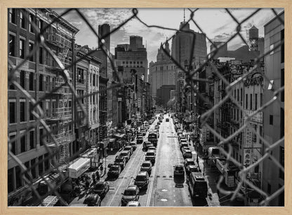 A high-angle, black and white photograph of a bustling New York City street viewed through a chain-link fence. The fence is in the foreground, framing the scene of traffic-filled streets, historic buildings with fire escapes, and skyscrapers in the distance under a cloudy sky. Wall Art