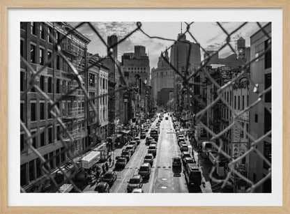 A high-angle, black and white photograph of a bustling New York City street viewed through a chain-link fence. The fence is in the foreground, framing the scene of traffic-filled streets, historic buildings with fire escapes, and skyscrapers in the distance under a cloudy sky. Wall Art