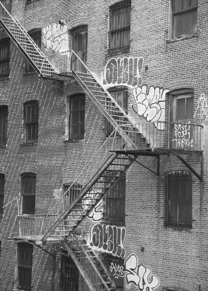 A black and white photograph of a brick apartment building with a prominent metal fire escape zigzagging across its facade. The walls are adorned with various styles of graffiti, and the windows are covered with bars, creating a gritty, urban scene. Decor
