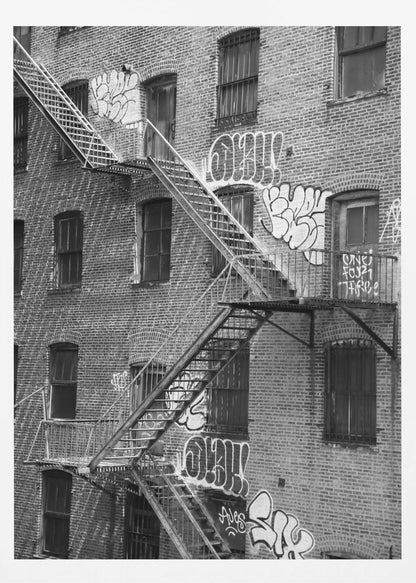 A black and white photograph of a brick apartment building with a prominent metal fire escape zigzagging across its facade. The walls are adorned with various styles of graffiti, and the windows are covered with bars, creating a gritty, urban scene. Decor