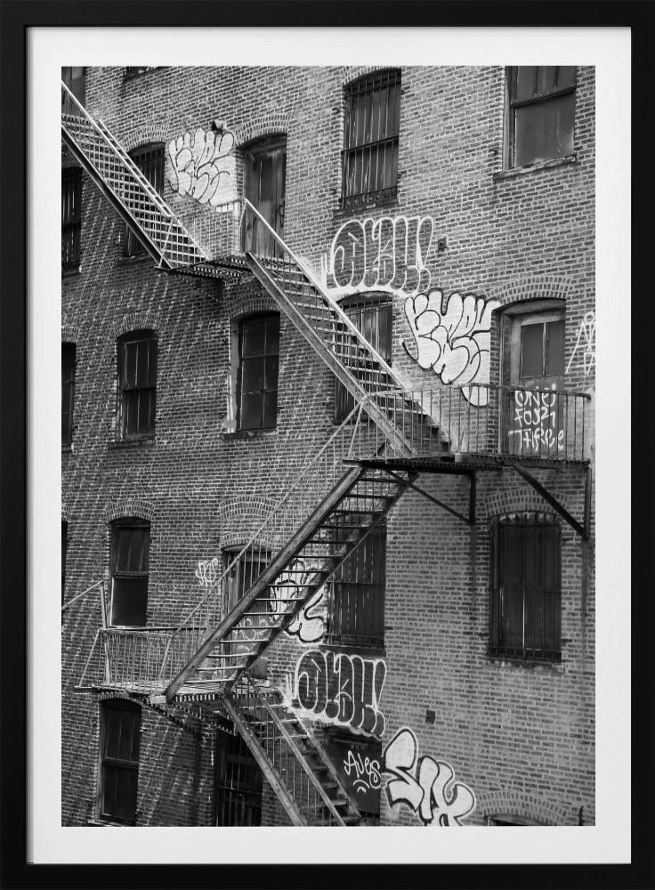 A black and white photograph of a brick apartment building with a prominent metal fire escape zigzagging across its facade. The walls are adorned with various styles of graffiti, and the windows are covered with bars, creating a gritty, urban scene. Decor
