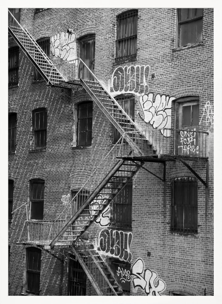 A black and white photograph of a brick apartment building with a prominent metal fire escape zigzagging across its facade. The walls are adorned with various styles of graffiti, and the windows are covered with bars, creating a gritty, urban scene. Decor