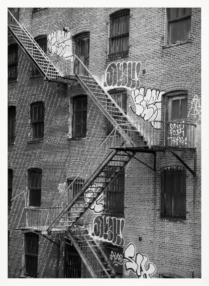 A black and white photograph of a brick apartment building with a prominent metal fire escape zigzagging across its facade. The walls are adorned with various styles of graffiti, and the windows are covered with bars, creating a gritty, urban scene. Decor