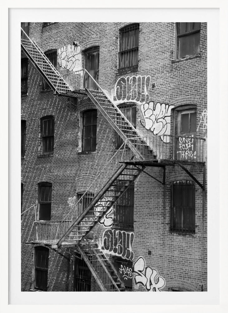 A black and white photograph of a brick apartment building with a prominent metal fire escape zigzagging across its facade. The walls are adorned with various styles of graffiti, and the windows are covered with bars, creating a gritty, urban scene. Decor